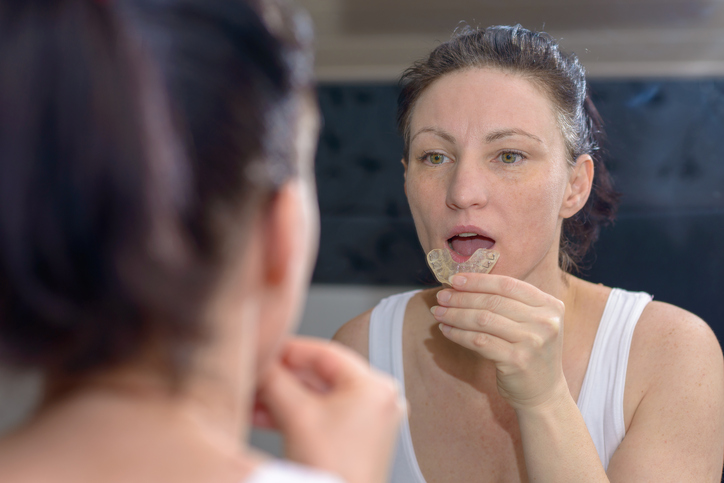 Woman holding an upper bite plate to prevent grinding her teeth together as she looks at her reflection in the mirror